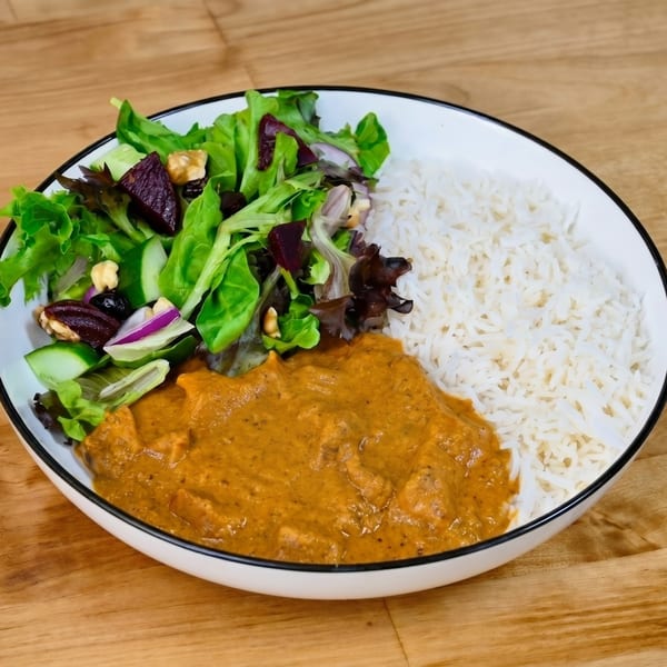 A white bowl filled with butter chicken, white rice, and a fresh green salad with walnuts.