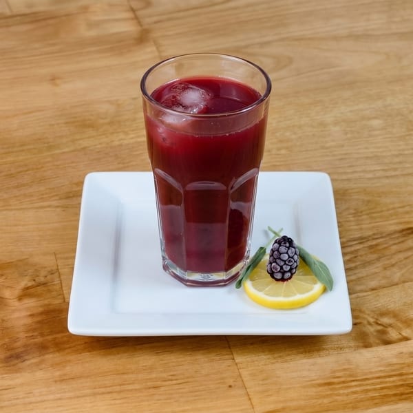 Dark red beverage with ice in a glass, garnished with a lemon slice, sage, and a blackberry.