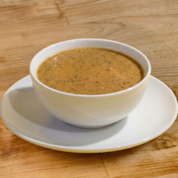 A white bowl of creamy, speckled brown soup sits on a white saucer on a wooden table.