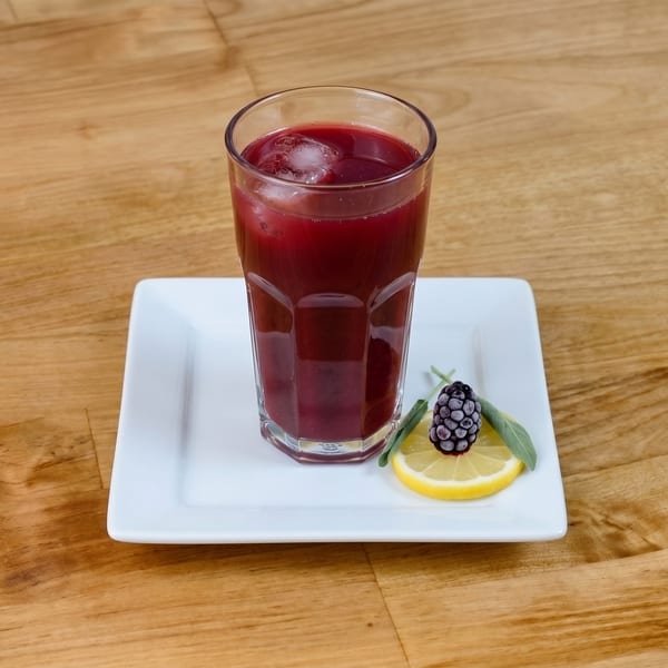 A glass of dark red juice with ice, garnished with a lemon slice, blackberry, and sage.