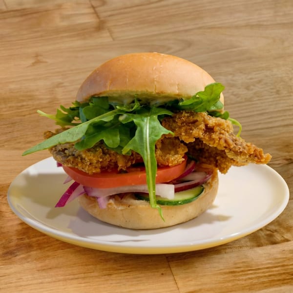 Crispy fried chicken sandwich with arugula, tomato, red onion, and cucumber on a white plate.